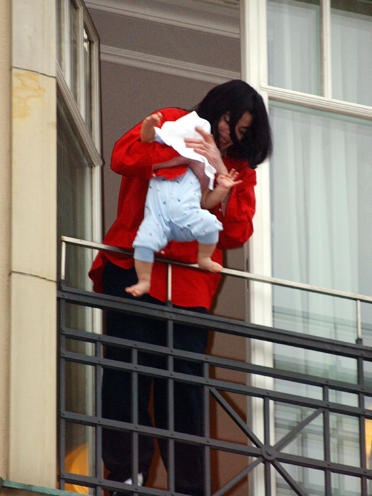 Michael Jackson într-o cămașă roșie în timp ce îl scoate pe Prince Michael 'Blanket' Jackson II pe un balcon în Berlin