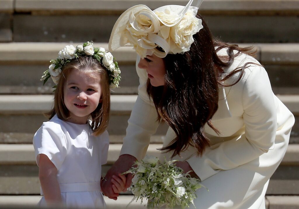 Prințesa Charlotte într-o rochiță albă în timp ce poartă pe cap o coroniță de flori, iar mama sa, Kate Middleton o ține de mână