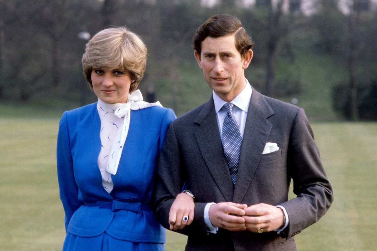 Prințesa Diana într-un costum albastru în timp ce se află lângă Prințul Charles care poartă un costum gri și zâmbesc la camera de fotografiat
