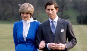 Prințesa Diana într-un costum albastru în timp ce se află lângă Prințul Charles care poartă un costum gri și zâmbesc la camera de fotografiat