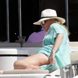 Actrița Rebel Wilson în timp ce stă întinsă și face plajă pe un iaht de lux, purtând o pălărie de plajă și o rohie lungă albastră în timp ce își descoperă un picior