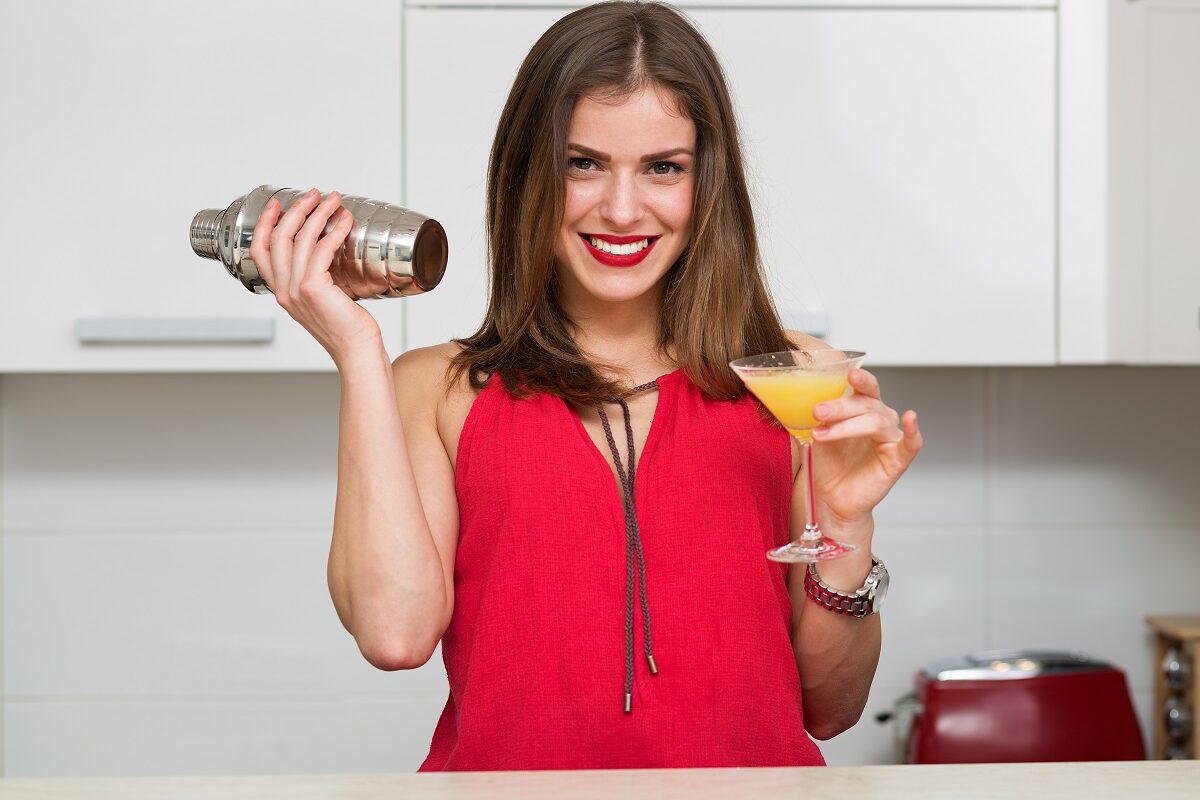 O femeie frumoasă care se află într-o bucătărie și ține în mână un pahar cu un cocktail fără alcool și un shaker