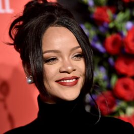 Artista Rihanna care a devenit miliardară purtând o rochie neagră pe gât și părul prins într-un coc lejer în timp ce zâmbește la cameră și se află pe covorul roșu