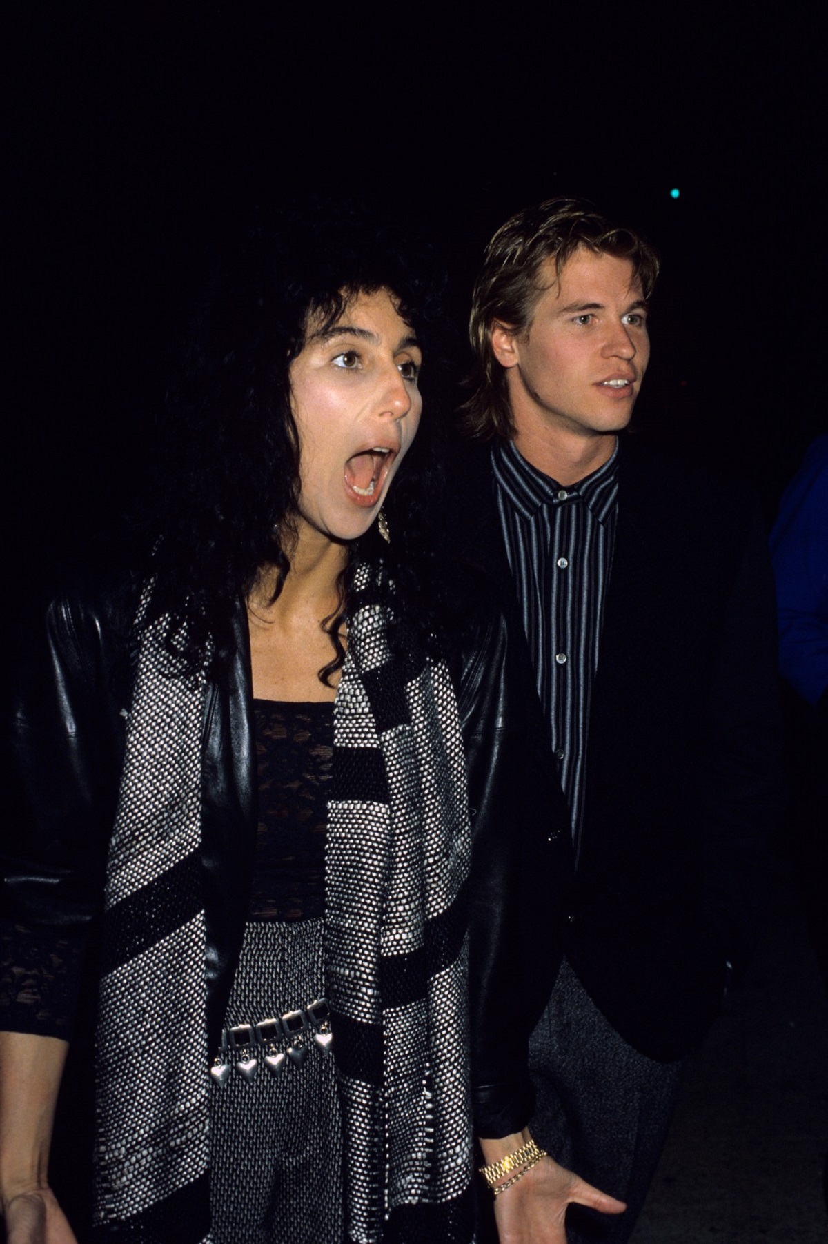Cher în timp ce strigă și poartă o fustă gri, o bluză neagră și un fular gri cu negru, alături de fostul său iubit Val Kilmer în anul 1984