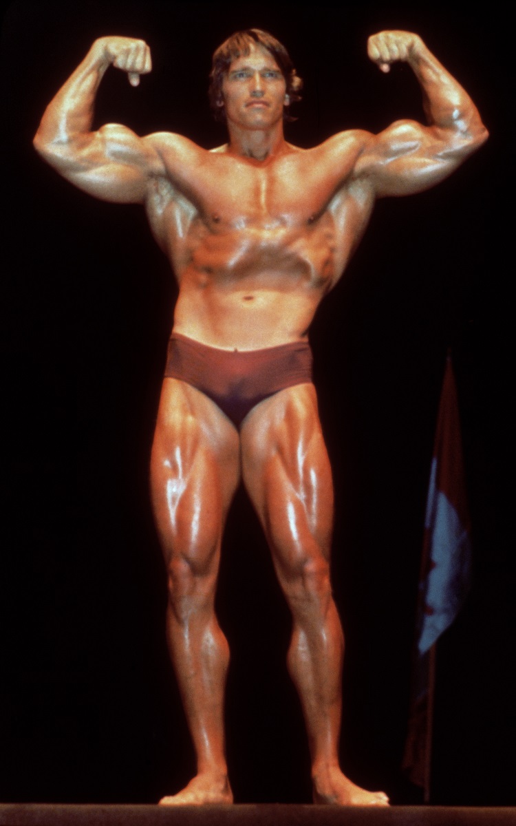 Arnold Schwarzenegger, la o competiție de bodybuilding din anii 1980. Mușchii mari, dați cu ulei, slip de culoare maro, fundal negru