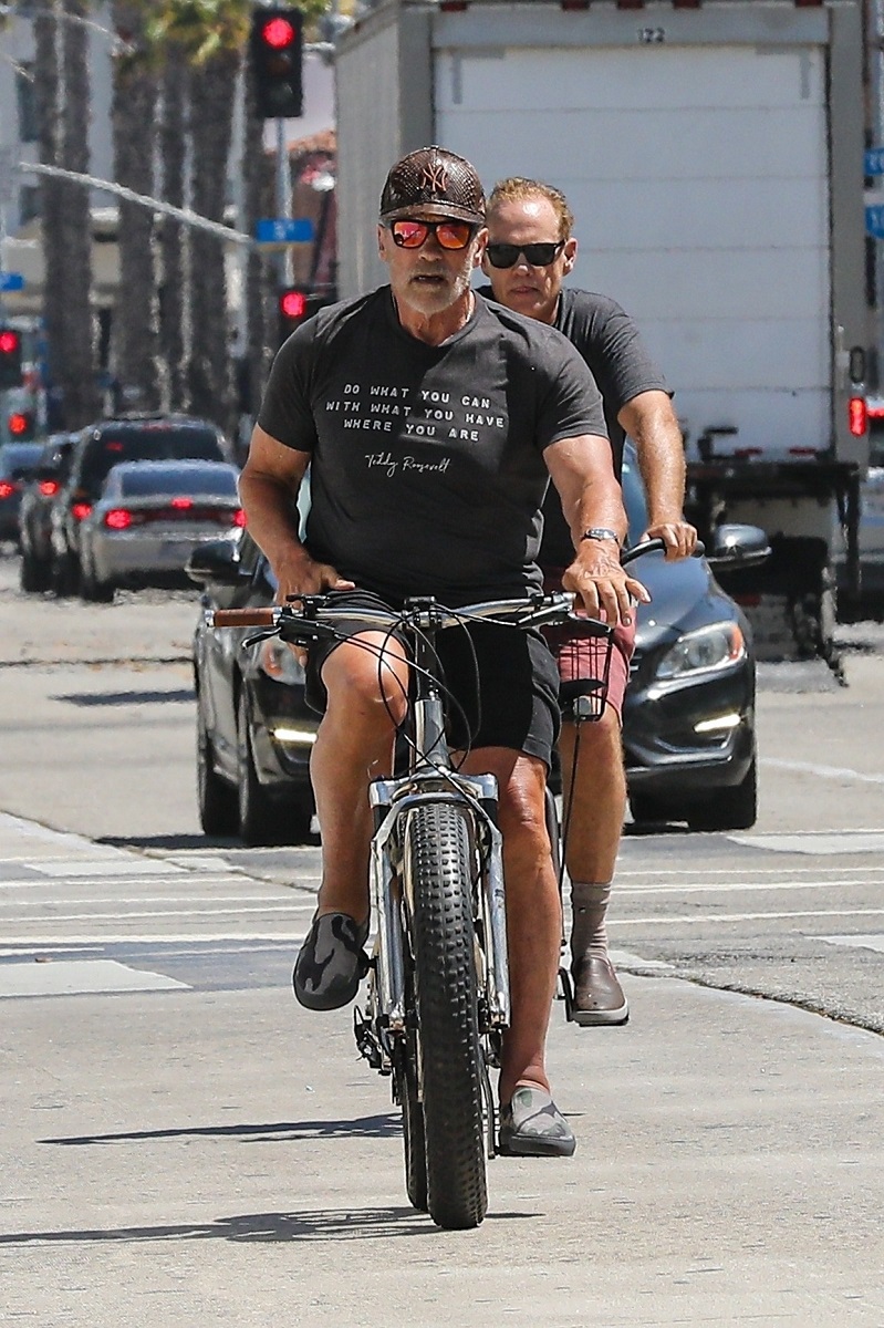Arnold Schwarzenegger pe bicicletă, în Santa Monica, California, anul 2021. El e îmbrăcat în negru, pe bicicletă, are un prieten în spate. Fundal cu stradă și mașini