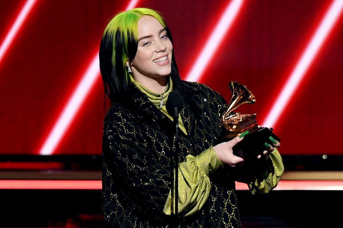 Billie Eilish, scena Premiilor Grammy, la cea de-a 62-a editie, în anul 2020. Eilish a purtat o bluză neagră deasupra, bluză verde dedesubt, are părul verde cu negru și un premiu Grammy în mână, cu fundal roșu