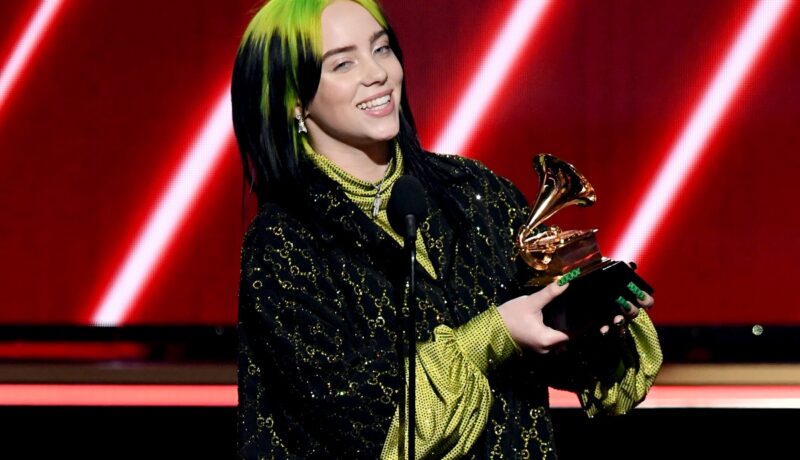 Billie Eilish, scena Premiilor Grammy, la cea de-a 62-a editie, în anul 2020. Eilish a purtat o bluză neagră deasupra, bluză verde dedesubt, are părul verde cu negru și un premiu Grammy în mână, cu fundal roșu