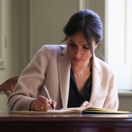 Meghan Markle a semnat cartea vizitatorilor în cadrul unei vizite la Casa Edes, Sussex, în anul 2018. Poartă o bluză neagră și o haină bej, cu fundal cu alb. Masa e din lemn maro