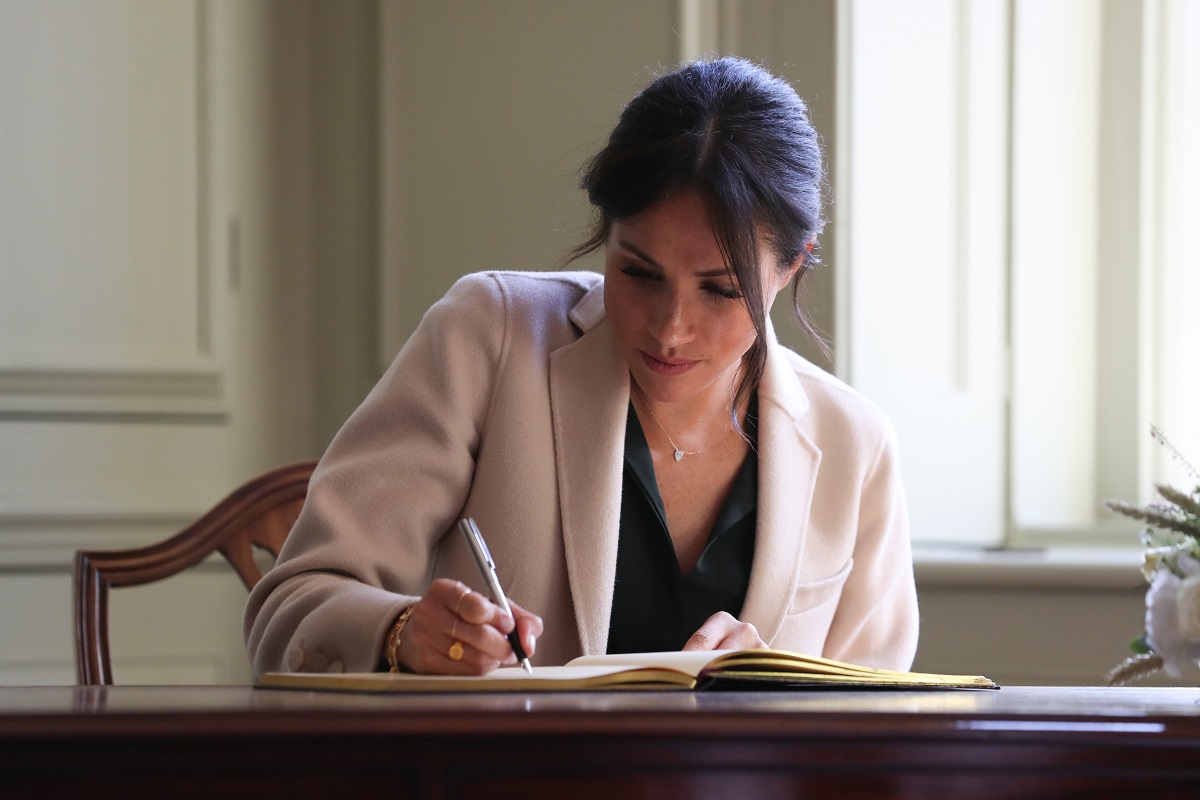 Meghan Markle a semnat cartea vizitatorilor în cadrul unei vizite la Casa Edes, Sussex, în anul 2018. Poartă o bluză neagră și o haină bej, cu fundal cu alb. Masa e din lemn maro