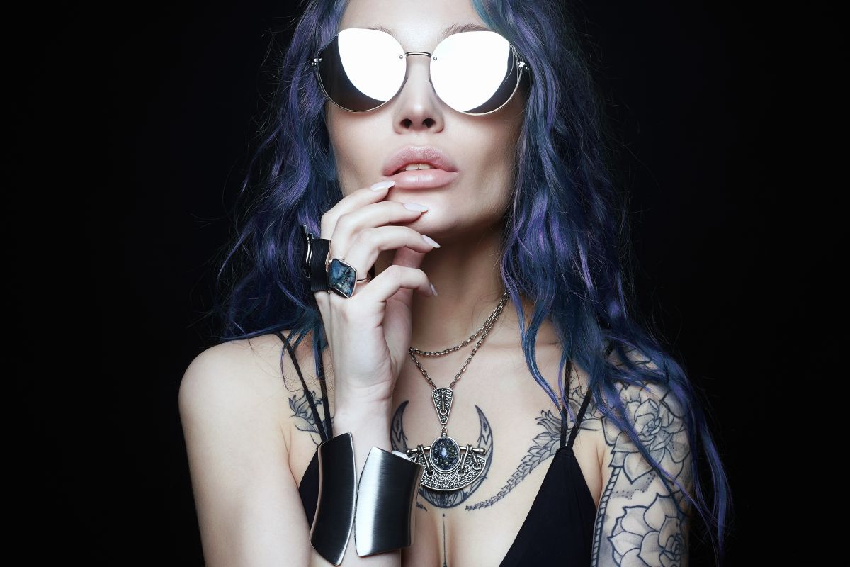 O femeie cu părul lung, albastru, care poartă ochelari de soare și bijuterii masive, are imprimate pe corp tatuaje.
