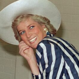 Prințesa Diana, într--un costum cu dungi albe și albastru închis, pălărie albă. Zâmbește și se uită la cameră, pe fundal e un perete bej