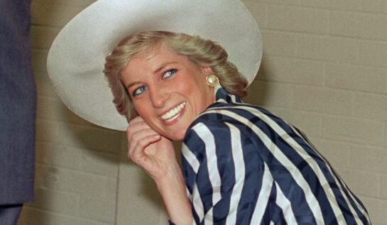 Prințesa Diana, într--un costum cu dungi albe și albastru închis, pălărie albă. Zâmbește și se uită la cameră, pe fundal e un perete bej