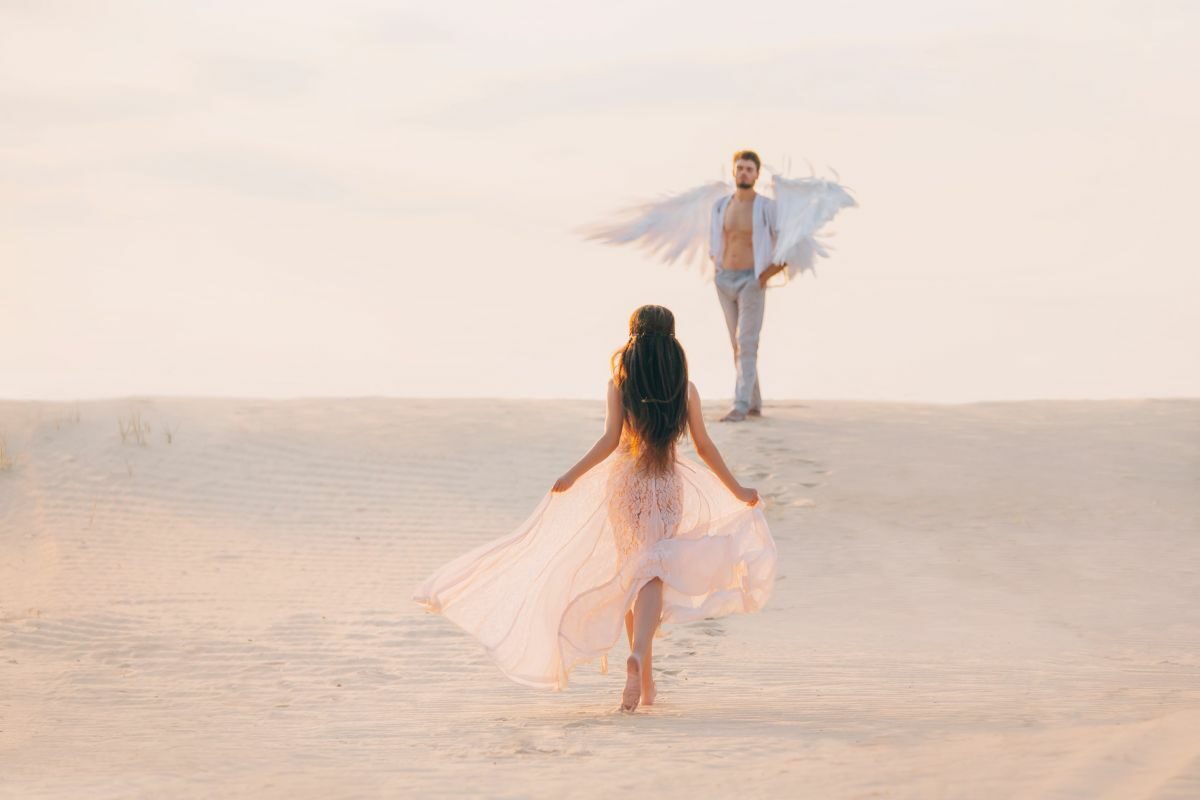 O femeie cu părul lung, îmbrăcată în alb, pășeste către un bărbat, care are aripi de îngeri.