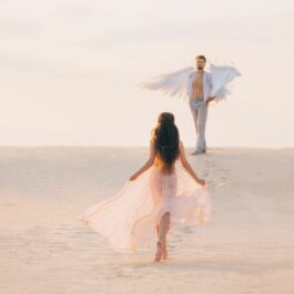 O femeie cu părul lung, îmbrăcată în alb, pășeste către un bărbat, care are aripi de îngeri.