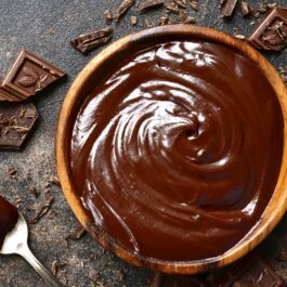 Cremă ganache de ciocolată într-un bol de lemn, alături de bucăți de ciocolată și o linguriță cu cremă
