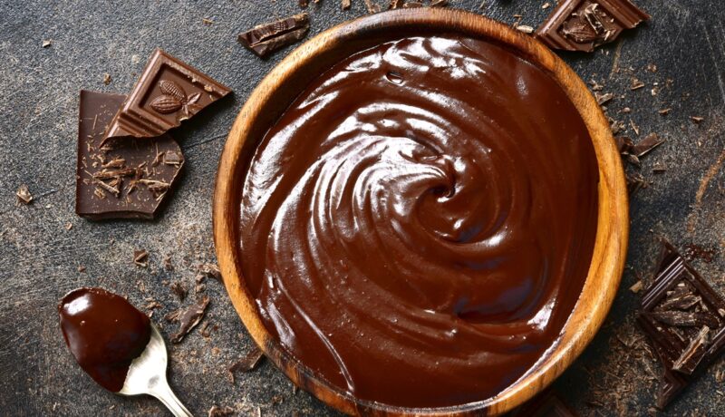 Cremă ganache de ciocolată într-un bol de lemn, alături de bucăți de ciocolată și o linguriță cu cremă