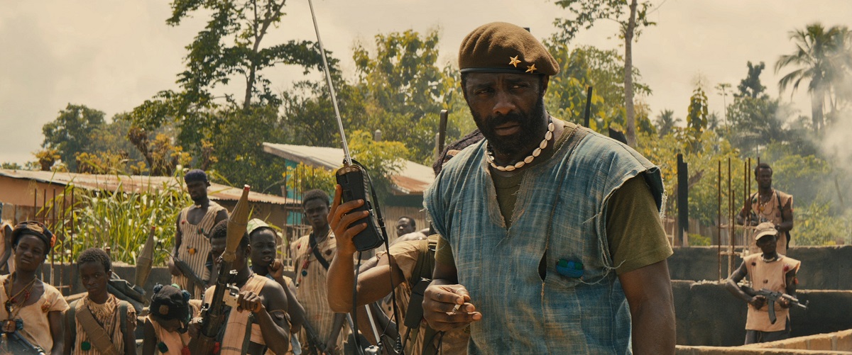 Idris Elba în filmul Beasts of No Nation. Poartă o bluză din blugi, cu decolteu, și o baretă. Fundal tropical cu război
