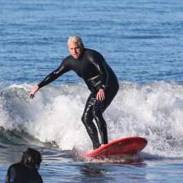 Jonah Hill la mare, în timp ce face surfing. Placa lui e roșie și costumul lui e negru, cu un val pe fundal