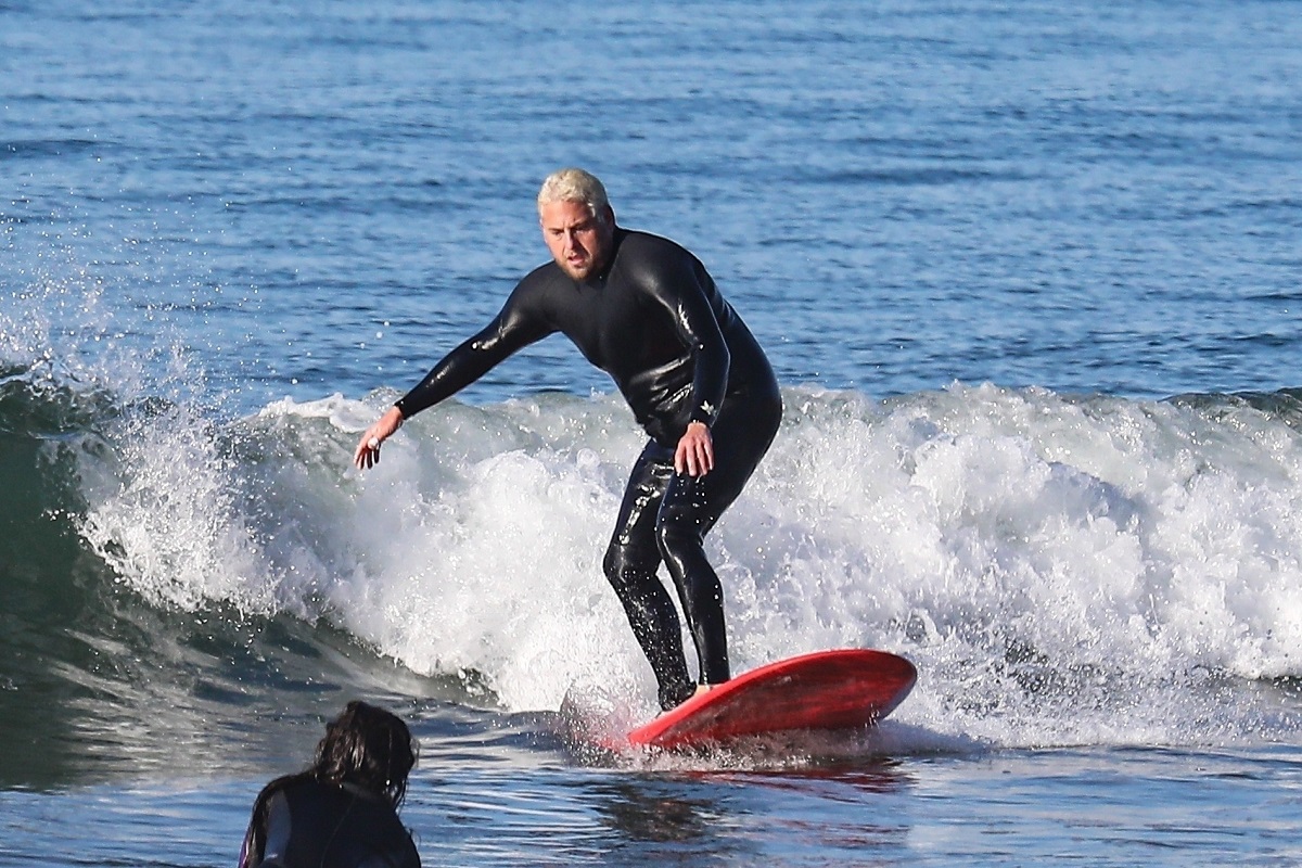 Jonah Hill la mare, în timp ce face surfing. Placa lui e roșie și costumul lui e negru, cu un val pe fundal