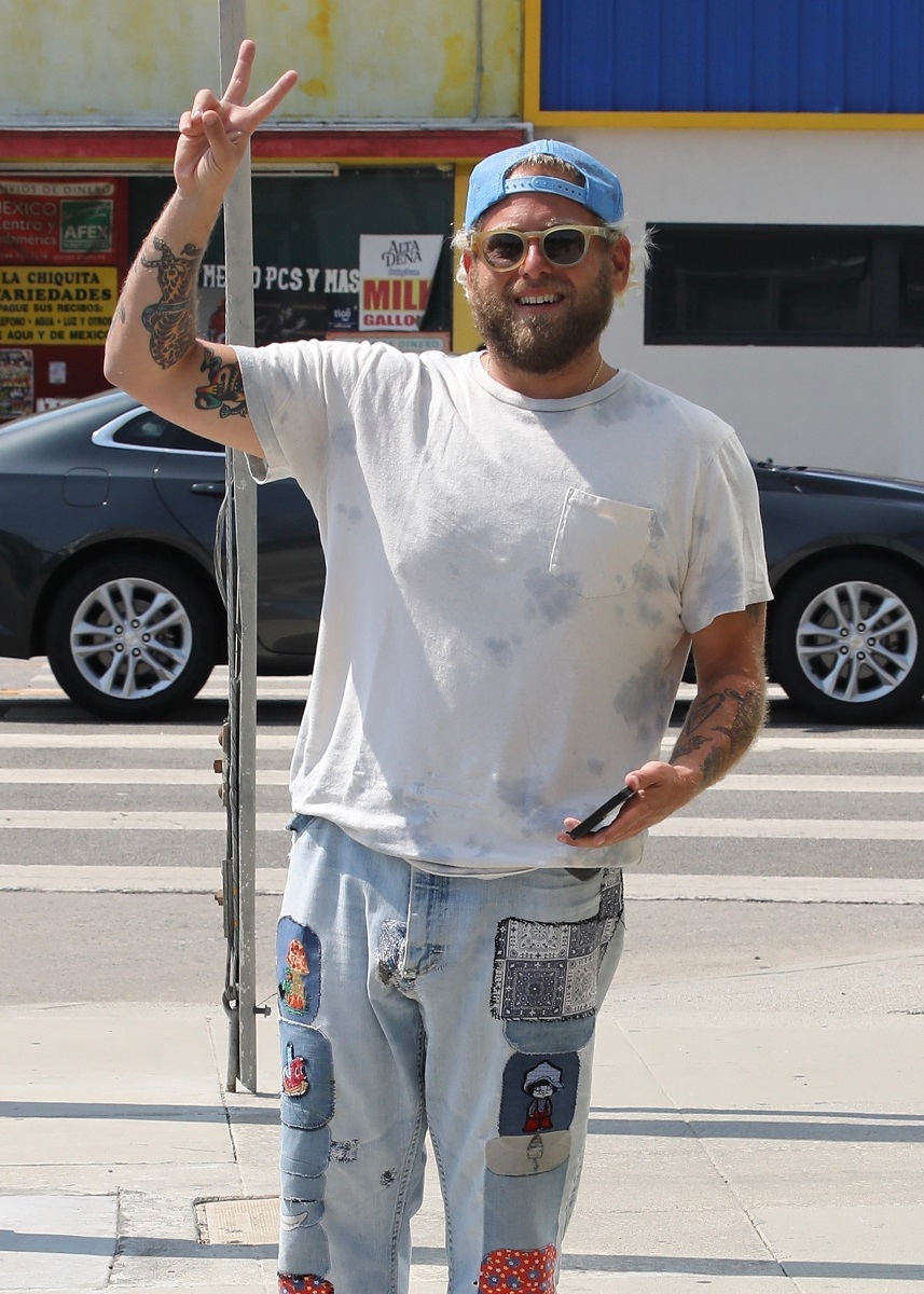 Jonah Hill surprins în Venice, California, în anul 2021, îmbrăcat în blugi deschiși la culoare, cu aplicații colorate, tricou alb, șapcă albastră și ochelari de soare. Strada pe fundal