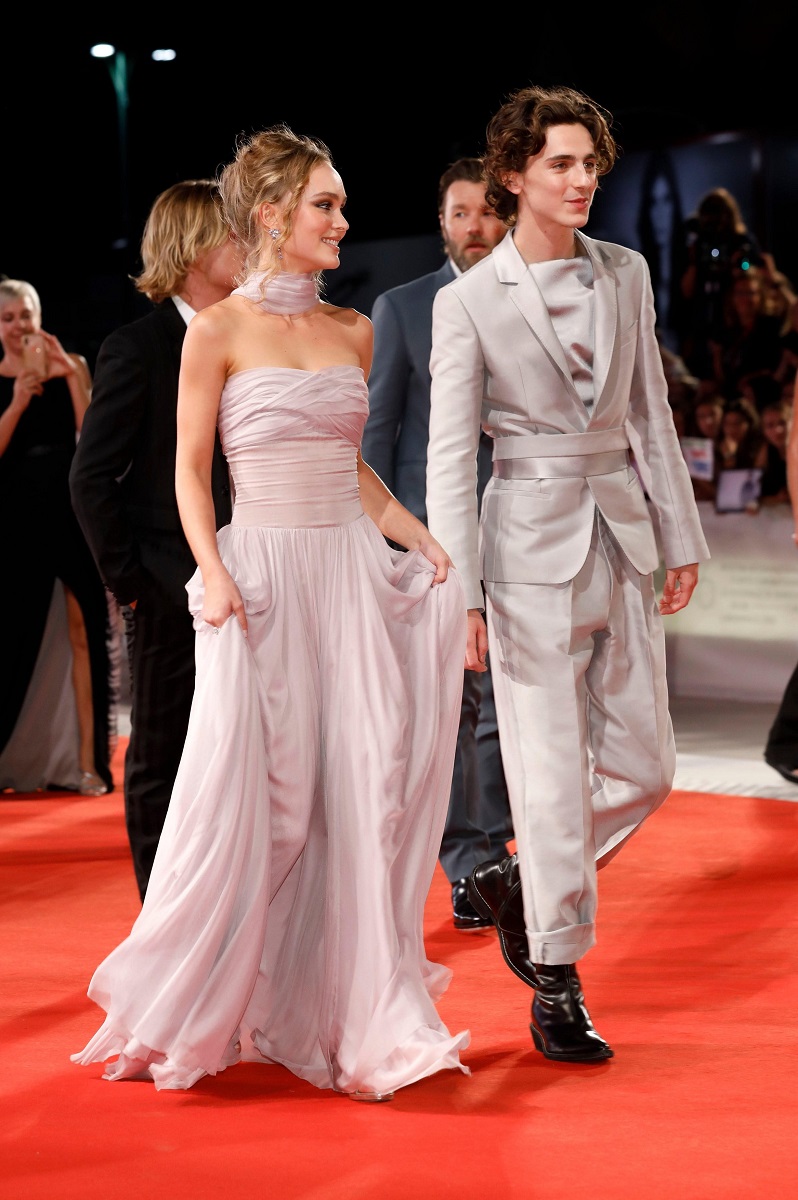 Lily-Rose Depp și Timothee Chalamet pe covorul roșu de la Veneția, 2019, la premiera filmului The King. El poartă un costum argintiu, prins în talie, ea poartă o rochie roz, lungă, fără bretele