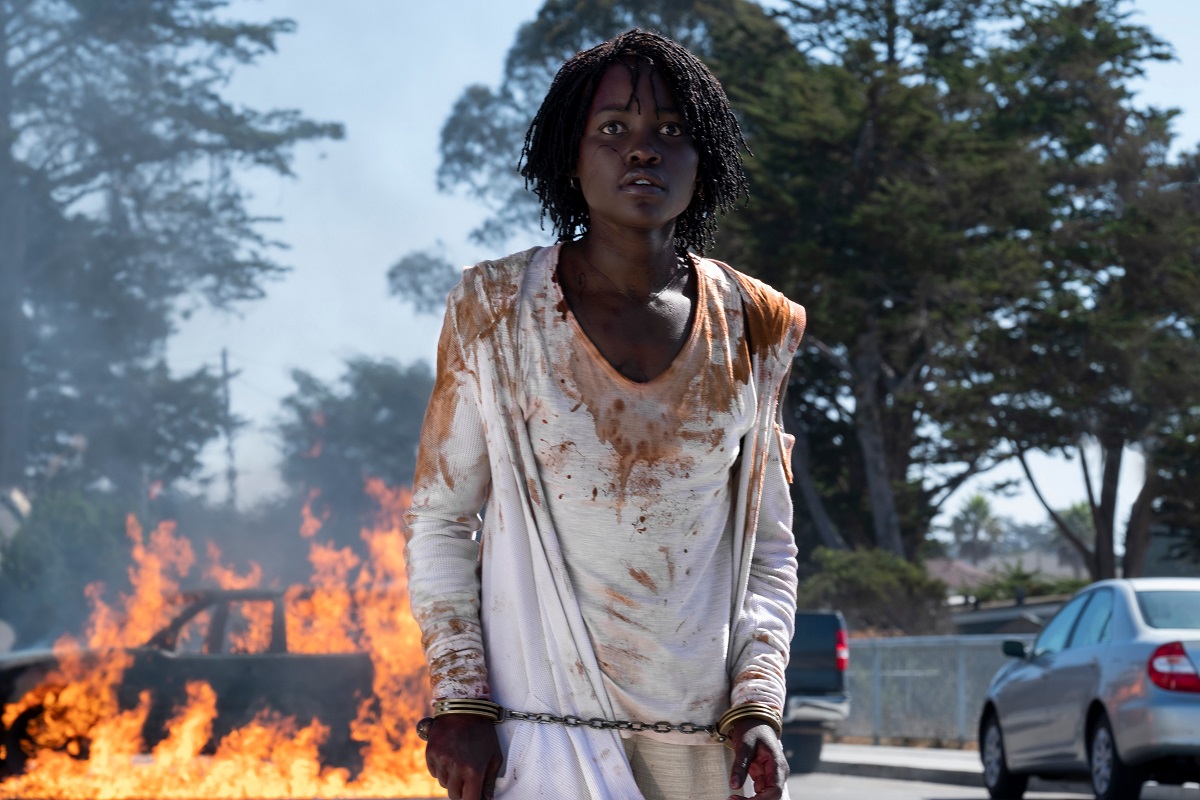 Lupita Nyong'o în filmul Us. E îmbrăcată în haine albe, murdărite de explozie. Foc și explozii pe fundal