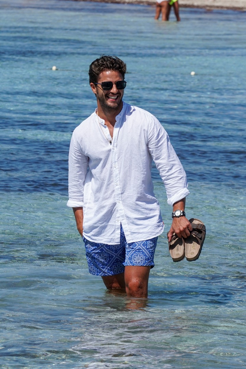Maxi Iglesias, în mare, cu o pereche de șlapi în mână, îmbrăcat într-o cămașă albă