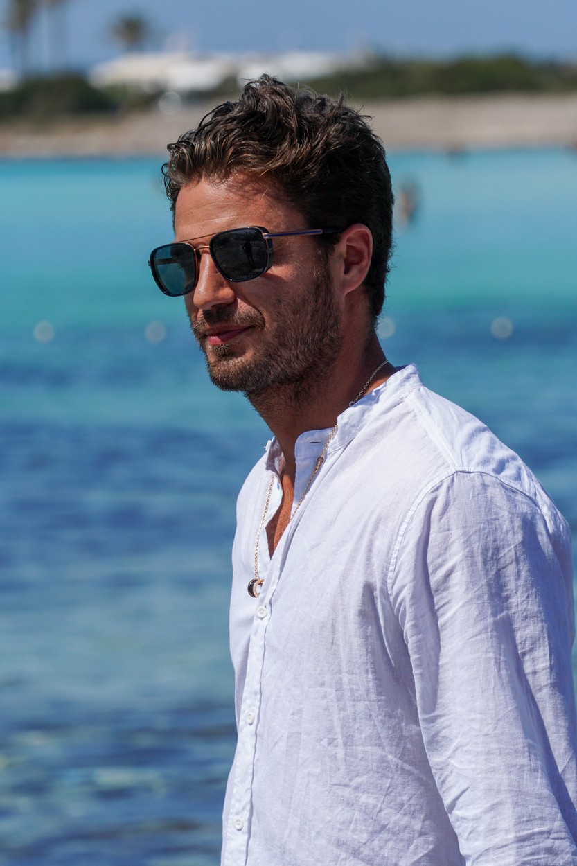 Maxi Iglesias, cu ochelari de soare, într-o cămașă albă, la plajă, în timpul unei ședințe foto