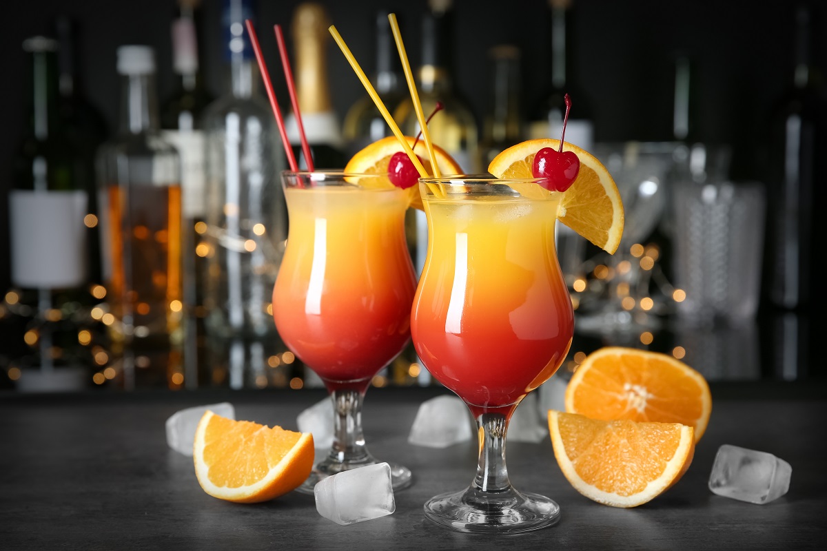 Două pahare care stau pe un bar umplute cu băutura cuddles on the beach lângă care se află bucăți de portocală