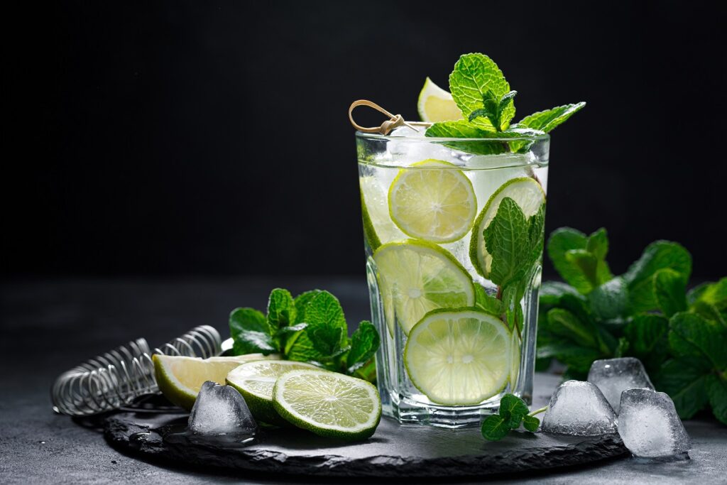 A tray filled with a cup of mojito garnished with mint along with several mint leaves, ice and lime