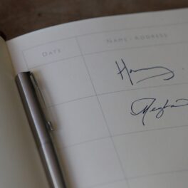 Semnătura lui Meghan Markle din cartea vizitatorilor la Casa Edes, în regiunea Sussex, din anul 2018. prințul Harry are prima semnătură, Meghan a doua, fundal alb, un pix în mijlocul cărții