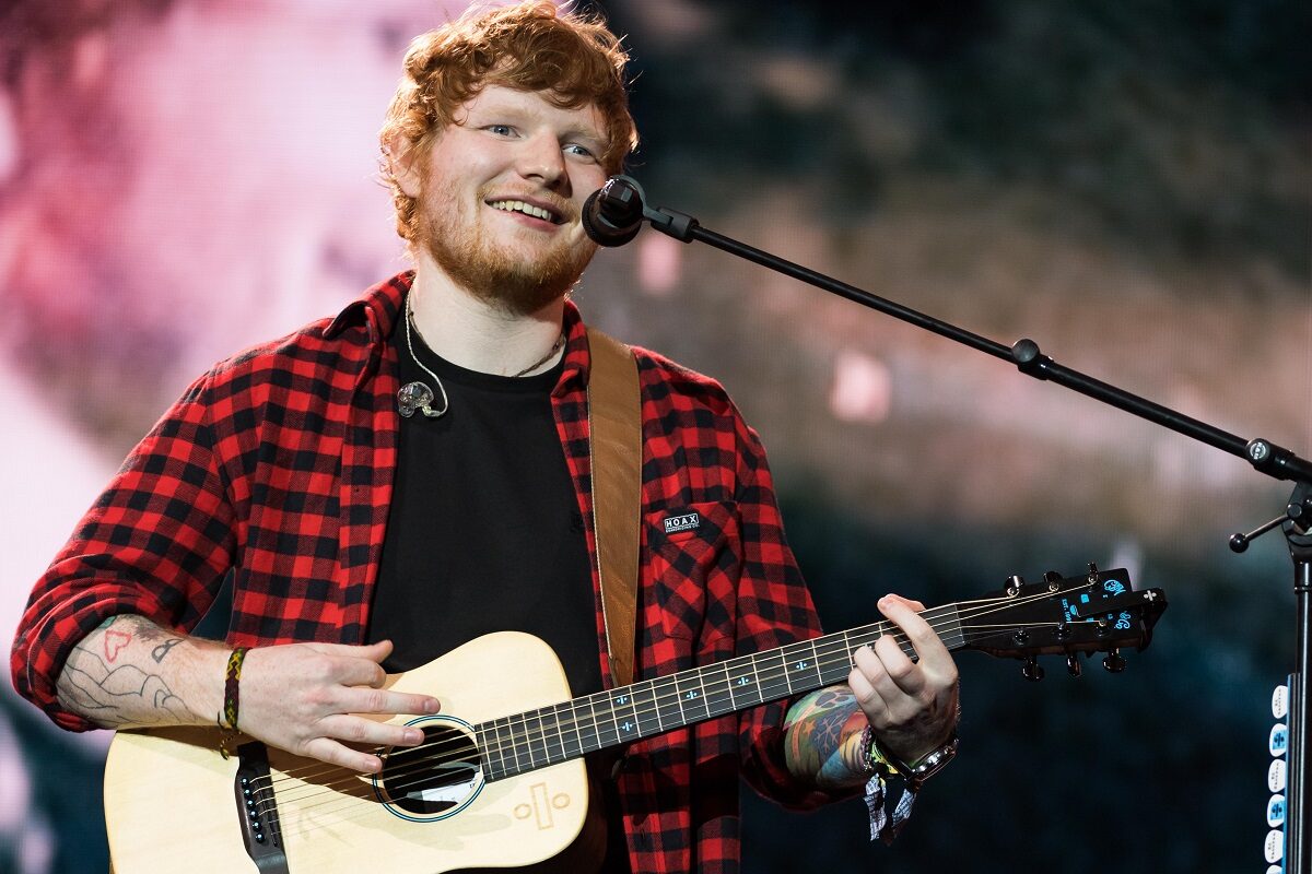 Ed Sheeran pe scenă purtând un tricou negru și o cămașă roșie în carouri în timp ce cântă la microfon în timpul unui concert