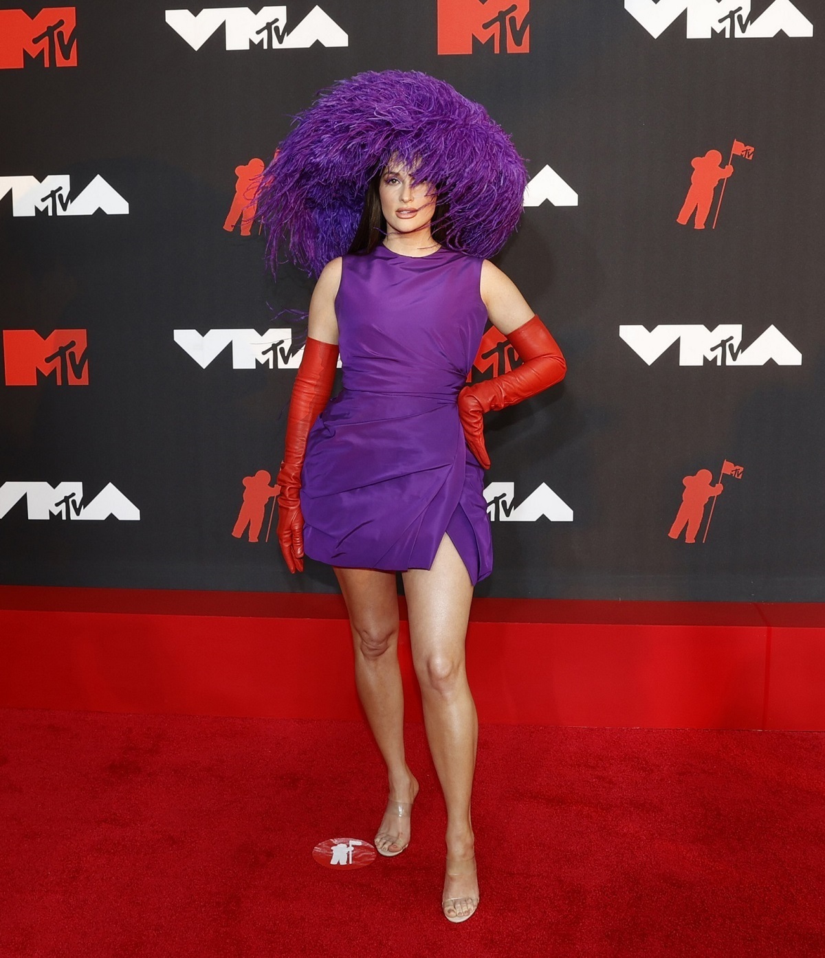 Kacey Musgraves într-o rochie mov cu o pălărie cu pene și o pereche de mănuși din latex roșii la MTV VMA 2021