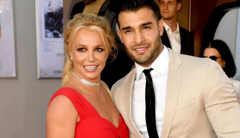 Britney Spears într-o rochie roșie alături de logodnicul său Sam Asghari care poartă un costum crem și o cravată neagră