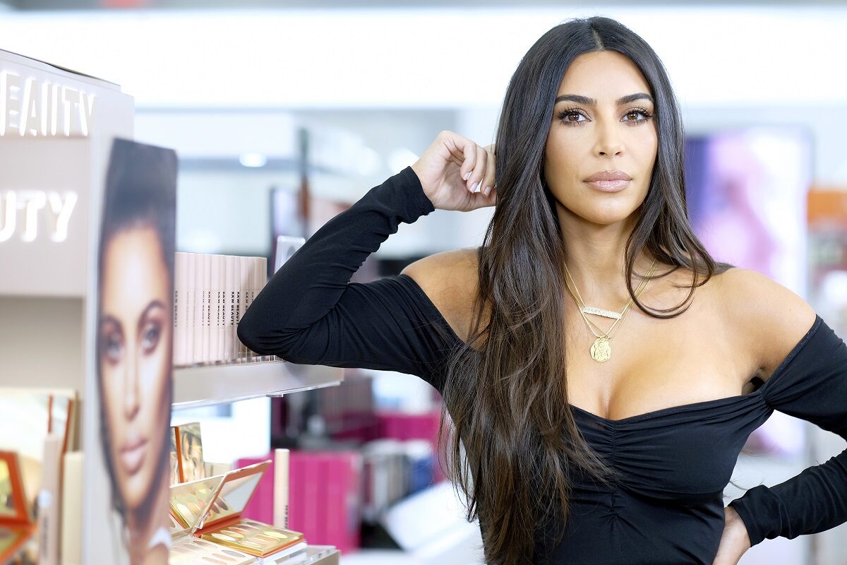 Machiajul lui Kim Kardashian natural care poartă o bluză neagră și stă sprijinită cu fața într-o mână la lansarea unui brand de machiaj