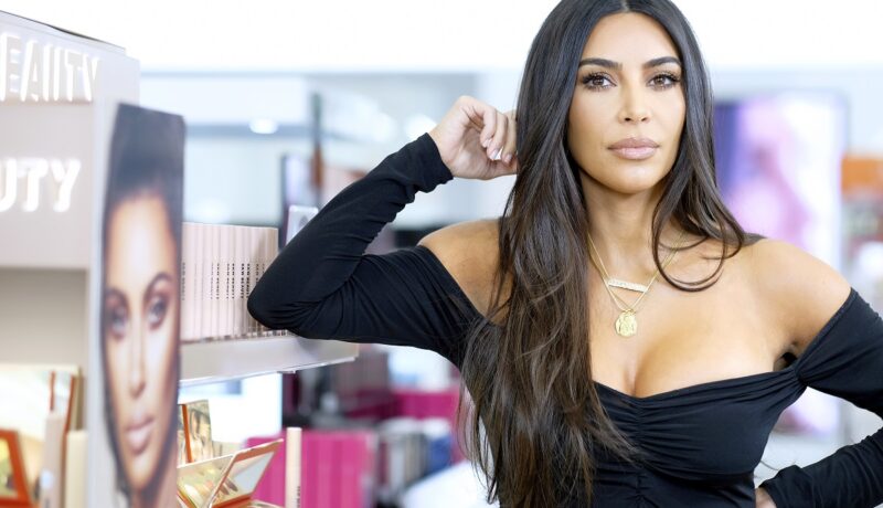 Machiajul lui Kim Kardashian natural care poartă o bluză neagră și stă sprijinită cu fața într-o mână la lansarea unui brand de machiaj
