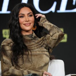 Kim Kardashian purtând o bluză maro în timp ce stă pe scaun și își așează părul după ureche