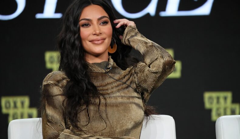 Kim Kardashian purtând o bluză maro în timp ce stă pe scaun și își așează părul după ureche