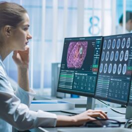 O femeie frumoasă îmbrăcată în halat alb de medic care stă în fața unui calculator și studiază mai multe imagini ale creierului pentru a-și da seama care sunt primele simptome ale demenței