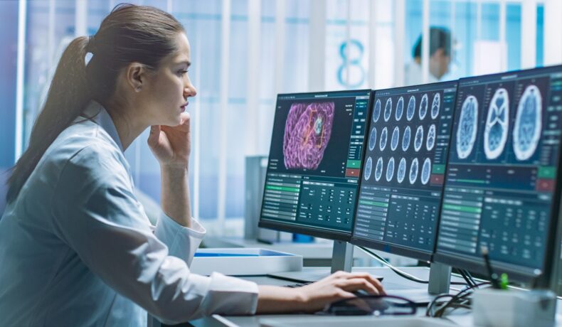 O femeie frumoasă îmbrăcată în halat alb de medic care stă în fața unui calculator și studiază mai multe imagini ale creierului pentru a-și da seama care sunt primele simptome ale demenței