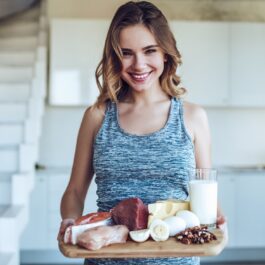 O femeie frumoasă cu parul blond care ține în mână o tavă cu un pahar de lapte, ouă și carne pentru a arăta care sunt acele semne ale deficienței de proteine din organism și ce alimente trebuie să consumăm pentru a le rezolva
