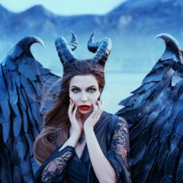 O femeie frumoasă care poartă aripi negre și o pereche de coarne în timp ce se mângâie pe față pentru a ilustra una din acele zodii invidioase