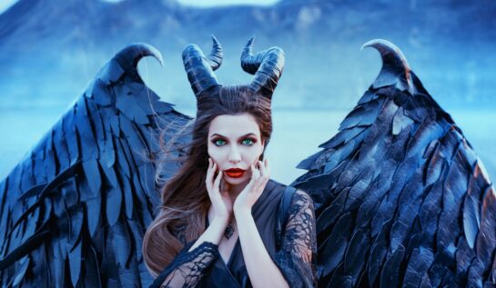 O femeie frumoasă care poartă aripi negre și o pereche de coarne în timp ce se mângâie pe față pentru a ilustra una din acele zodii invidioase