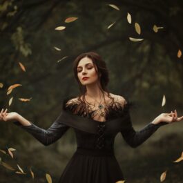 O femeie frumoasă îmbrăcată într-o rochie neagră în timp ce se află într-o pădure iar în jurul său plutesc în cerc frunzele copacilor pentru a potrtretiza una din acele zodii răzbunătoare
