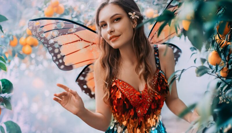 O femeie frumoasă cu aripi de fluture care poartă un top portocaliu și o fustă albastră în timp ce face cu mâna printre flori pentru a reprezenta una din acele zodii vorbărețe