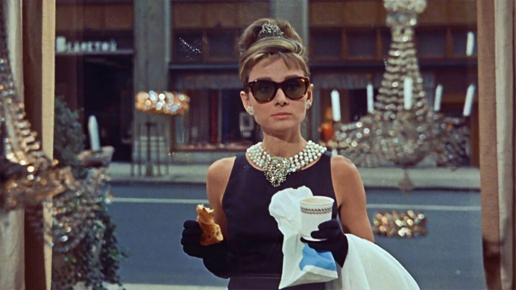 Audrey Hepburn în filmul Breakfast at Tifannys. Poartă celebra rochie neagră, cu perle și ochelari de soare. Stă în fața vitrinei magazinului și mănâncă