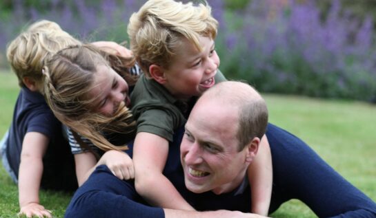 Prințul William se joacă alături de Prințul George, Prințesa Charlotte și prințul Louis. Toți trei sunt îmbrăcați în albastru închis și se joacă pe iarbă