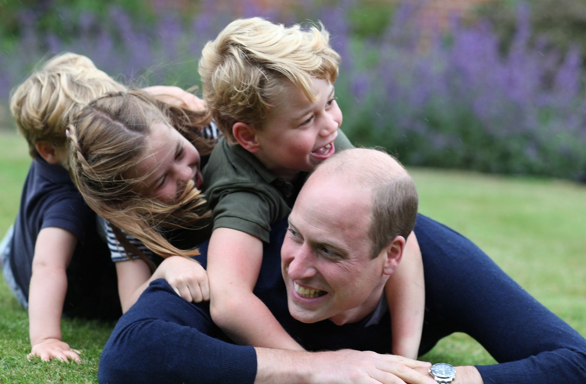 Prințul William se joacă alături de Prințul George, Prințesa Charlotte și prințul Louis. Toți trei sunt îmbrăcați în albastru închis și se joacă pe iarbă