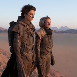Film still cu Timothee Chalamet și Rebecca Ferguson, din filmul Dune, 2021. Ei sunt îmbrăcați în armuri negre, cu un fundal cu deșert